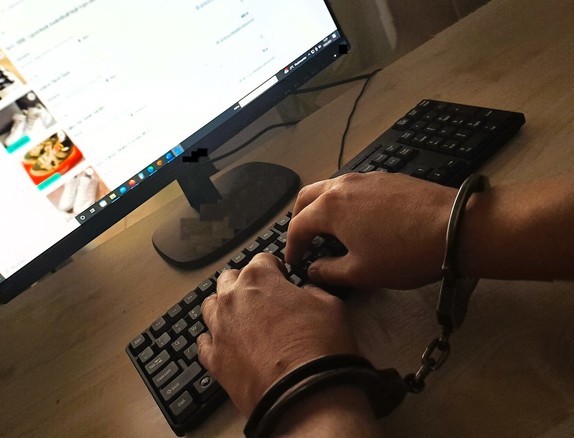 Ręce z złożonymi kajdankami nad klawiatura komputera w tle monitor