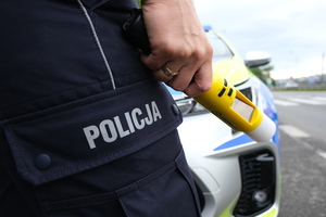 Policjant trzymający alkomat w tle radiowóz. Policjant widoczny w części - nogi - spodnie z napisem policja.