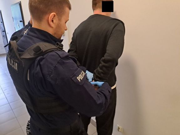 Zatrzymany mężczyzna ma zakładane kajdanki na ręce trzymane z tyłu przez umundurowanych funkcjonariuszy policji w pomieszczeniu dla osób zatrzymanych