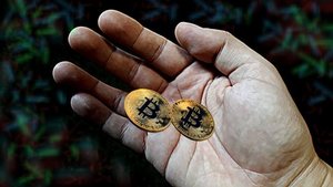 Dwie monety koloru złotego z logiem kryptowaluty Bitcoin trzymany w dłoni. Tło jest koloru ciemnego w górnym lewym rogu jest logo policji lubelskiej