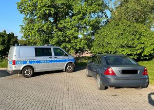 Radiowóz policyjny bus  oraz  szary sedan marki ford stojące na placu utwardzonym kostką brukową. W tle drzewa i zarośla.