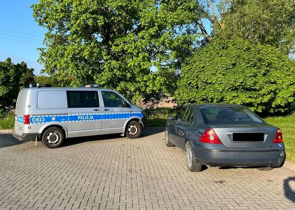 Radiowóz policyjny bus  oraz  szary sedan marki ford stojące na placu utwardzonym kostką brukową. W tle drzewa i zarośla.
