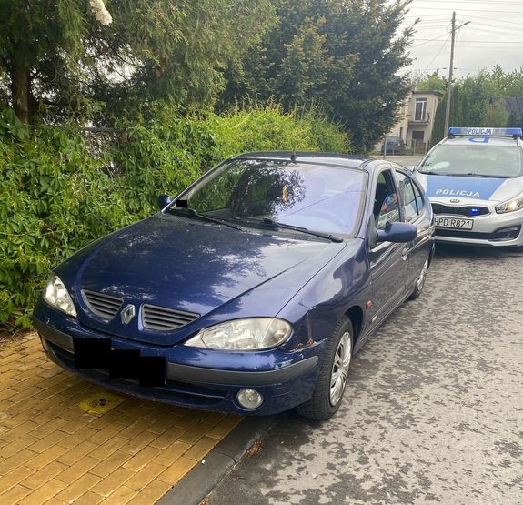 samochód osobowy koloru granatowego z uszkodzonym tylnym zawieszeniem stojący na drodze - częściowo na chodniku i jezdni. W tle oznakowany radiowóz policji z sygnałami włączonymi marki KIA