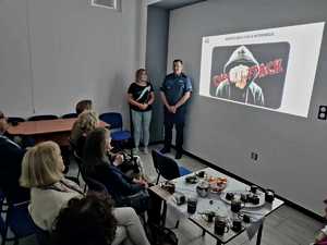 Uczestnicy spotkania w bibliotece w Dęblin. Przy wyświetlanej na ścianie prezentacji stoi umundurowany policjant. Goście siedzą na krzesłach