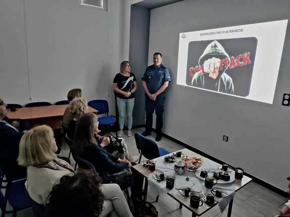 Uczestnicy spotkania w bibliotece w Dęblin. Przy wyświetlanej na ścianie prezentacji stoi umundurowany policjant. Goście siedzą na krzesłach