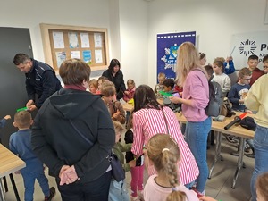 Grupa kilkunastu dzieci podczas zwiedzania budynku Komendy Powiatowej Policji w Rykach w sali odpraw. Policjant rozdaje odblaski