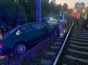 Uszkodzony po zdarzeniu samochód osobowy stojący na nasypie kolejowym przy słupie w tle pociąg oraz przedstawiciele służb