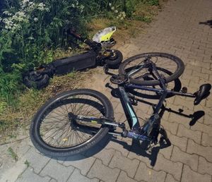 Rower i hulajnoga elektryczna leżące na na drodze rowerowej wyłożonej kostką brukową. w tle latarnie a na poboczu zarośla.