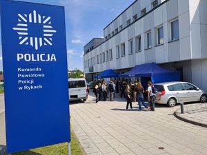 Zdjęcie stoiska policji ustawione przed budynkiem komendy policji w Rykach. Przy stoisku są odwiedzający oraz stoją pojazdy policyjne.