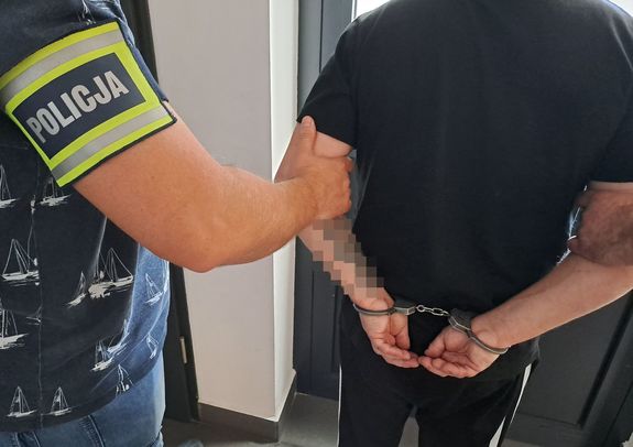 Policjant trzymający zatrzymanego mężczyznę za rękę . Zatrzymany ma zało0ózne kajdanki na ręce trzymane z tyłu i czarną koszulkę.