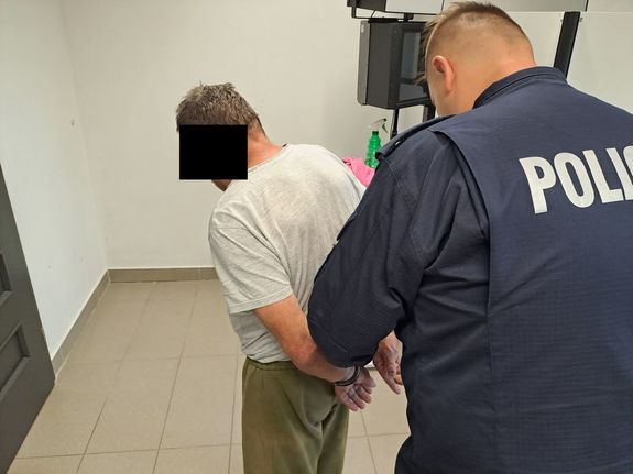 Policjant zakłada kajdanki na ręce trzymane z tyłu zatrzymanemu mężczyźnie w pomierzeniu policji