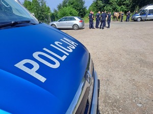Policjanci przygotowujący się do działań poszukiwawczych za zaginionym. widoczne są radiowozy policji zaparkowane na parkingu.