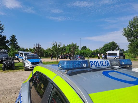 Policjanci przygotowujący się do działań poszukiwawczych za zaginionym. widoczne są radiowozy policji zaparkowane na parkingu.