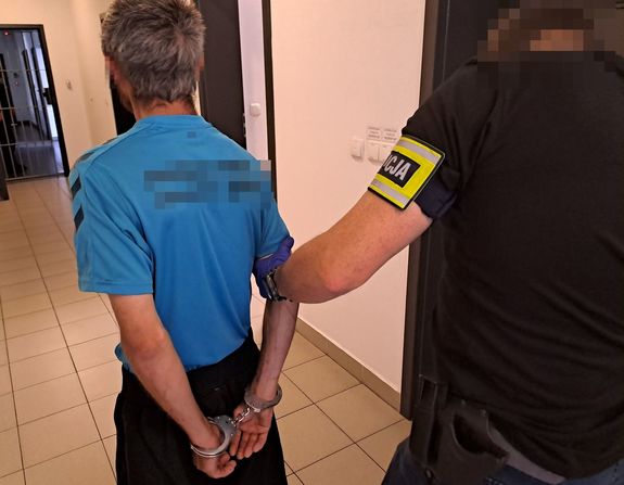 Zatrzymany mężczyzna z kajdankami założonymi na ręce z tyłu trzymany za rękę przez policjanta  nieumundurowanego z opaska z napisem policja