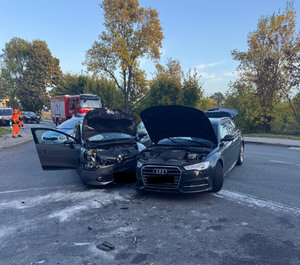 Dwa uszkodzone pojazdy w wyniku zdarzenia drogowego stojące na jedni. w tle pojazdy służb ratunkowych i porządkowych
