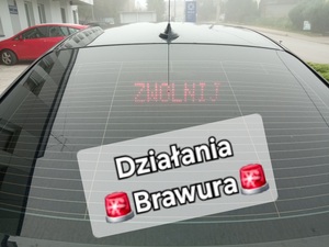 Tylna szyba radiowozów nieznakowany z wyświetlonym napisem zwolnij i dodany teks Działania brawura w tle budynek komendy policji w Rykach