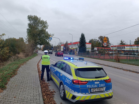 Policjant podczas nadzoru nad przejściem dla pieszych stoi obok oznakowanego radiowozu w tle droga pojazdy i szkoła