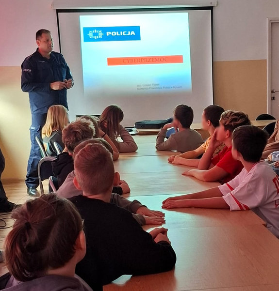 Policjant prowadzi spotkanie profilaktyczne dzieci spędzana krzesłach przy ławkach szkolnych w sali lekcyjnej i patrzą w kierunku policjant i wyświetlanego slajdu na ekranie