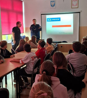 Policjant prowadzi spotkanie profilaktyczne dzieci spędzana krzesłach przy ławkach szkolnych w sali lekcyjnej i patrzą w kierunku policjant i wyświetlanego slajdu na ekranie