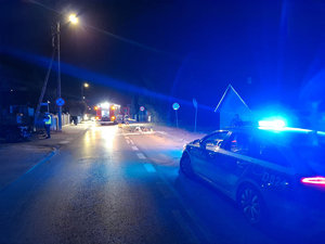 Miejsce wypadku drogowego w porze nocnej. Na ulicy leży uszkodzony motorower w tle pojazdy służb i funkcjonariusze wykonujący czynności służbowe.