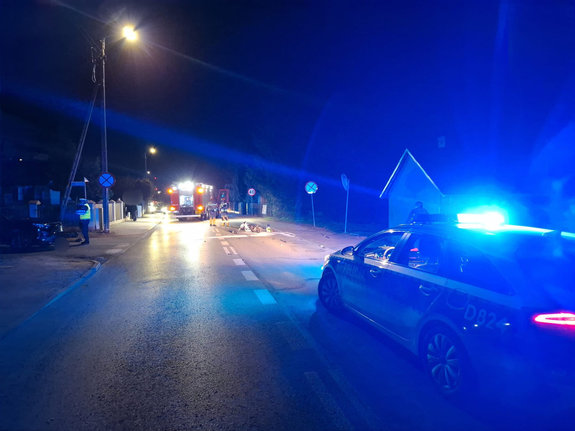 Miejsce wypadku drogowego w porze nocnej. Na ulicy leży uszkodzony motorower w tle pojazdy służb i funkcjonariusze wykonujący czynności służbowe.