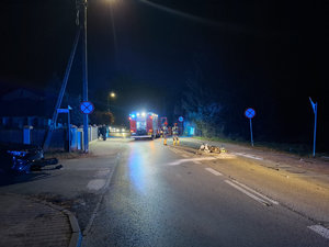Miejsce wypadku drogowego w porze nocnej. Na ulicy leży uszkodzony motorower w tle pojazdy służb i funkcjonariusze wykonujący czynności służbowe.