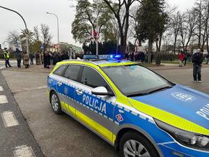 Powiatowe uroczystości w Rykach z okazji Narodowego Święta Niepodległości. Na miejscu w tle widać zgromadzone osoby a na pierwszym planie stoi radiowóz policji koloru srebrnego z włączonymi sygnałami barwy niebieskiej