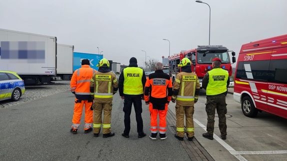 umundurowani funkcjonariusze służb stoją tyłem. W tle znajdują się samochody ciężarowe, radiowóz policyjny i wozy strażackie