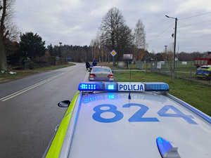 dach radiowozu oznakowanego z włączonymi sygnałami stojącego na poboczu przed nim stoi samochód osobowy Saab i kolejny radiowóz w tle zabudowa