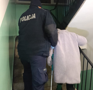 Umundurowany policjant prowadzący seniorkę z kulą na klatce schodowej
