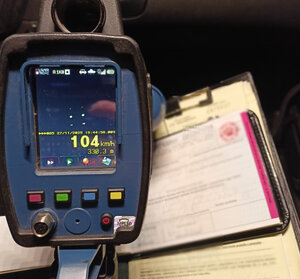 Zdjęcie miernika prędkości na którego ekranie wyświetlony jest pomiar pojazdu jadącego z prędkości 104 km/h w tle dokumenty - mandat  - zabudowany i wnętrze pojazdu