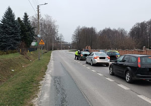Policjant podczas kontroli stanu trzeźwości kierującego na drodze stoją 3 samochody osobowe w tle radiowóz i inne zaparkowane pojazdy