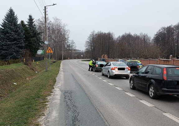 Policjant podczas kontroli stanu trzeźwości kierującego na drodze stoją 3 samochody osobowe w tle radiowóz i inne zaparkowane pojazdy
