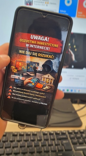 Telefon trzymany w ręku z wyświetloną grafiką ostrzegająca przed oszustwami w tle monitor komputera i klawiatura