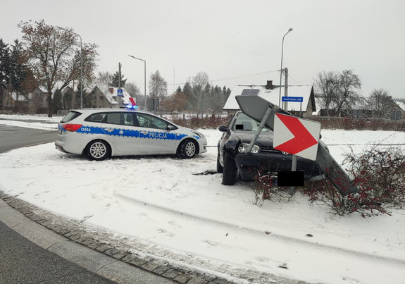 Uszkodzony Volkswagen zawieszony na znakach drogowych na rondzie obok stoi oznakowany radiowóz. teren przykryty śniegiem.