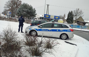 Uszkodzony Volkswagen zawieszony na znakach drogowych na rondzie obok stoi oznakowany radiowóz i policjant. teren przykryty śniegiem.