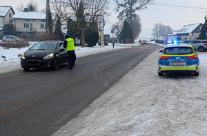 Policjant kontroluje stan trzeźwości osoby kierującej osobowym peugeotem koloru ciemnego w tle zabudowania a obok jezdni stoi oznakowany radiowóz w łączonymi sygnałami świetlnymi