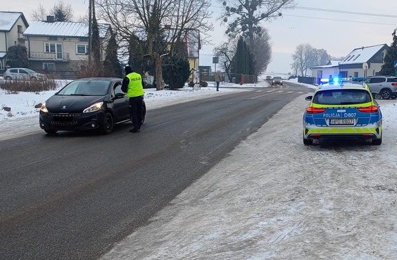 Policjant kontroluje stan trzeźwości osoby kierującej osobowym peugeotem koloru ciemnego w tle zabudowania a obok jezdni stoi oznakowany radiowóz w łączonymi sygnałami świetlnymi