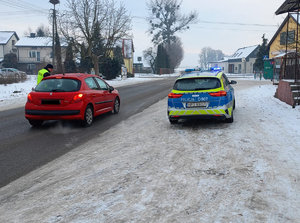 policjant kontroluje stan trzeźwości osoby kierującej osobowym peugeotem koloru czerwonego w tle zabudowania a obok jezdni stoi oznakowany radiowóz w łączonymi sygnałami świetlnymi