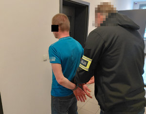 Zatrzymany mężczyzna w niebiskiej koszulce ma zakładane kajdanki na ręce z tyłu