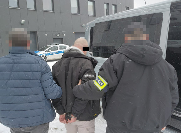 Dwóch policjantów pionu kryminalnego prowadzi zatrzymanego mężczyznę z kajdankami założonymi na ręce z tyłu.