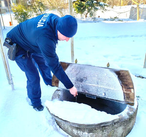Dzielnicowy nabiera wodę ze studni trzymając łańcuch w ręku, teren do około pokryty śniegiem.