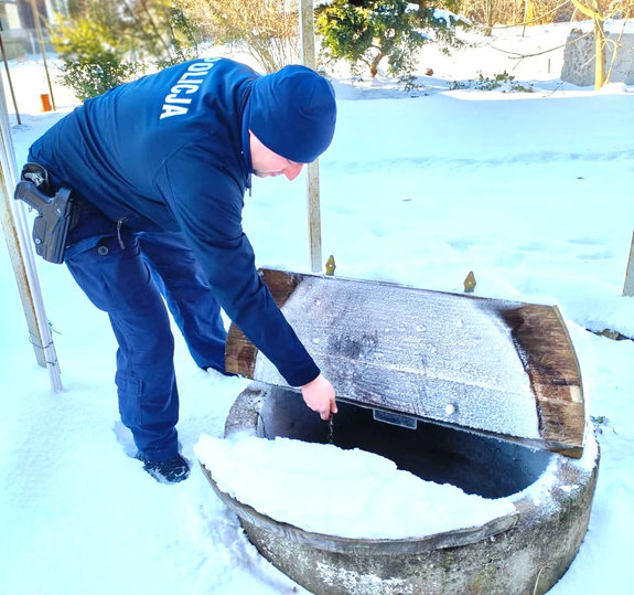 Dzielnicowy nabiera wodę ze studni trzymając łańcuch w ręku, teren do około pokryty śniegiem.