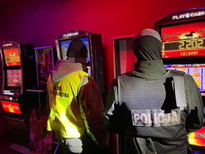 Zdjęcia wykonane wewnątrz lokalu w którym odbywały się gry hazardowe. Dwóch funkcjonariuszy policji i służby celno-skarbowej stoją przed automatami do gier.