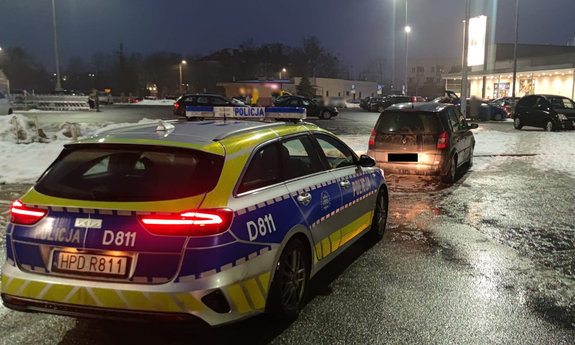 Radiowóz policji stoi na parkingu. Przed nim stoi osobowe renault w tle inne pojazdy zaparkowane oraz zabudowania