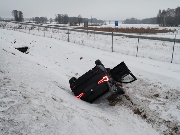 Pojazd marki BMW koloru Czernego  leżący w rowie. Obok stoi radiowóz policji i wystawiony jest znak uwaga wypadek