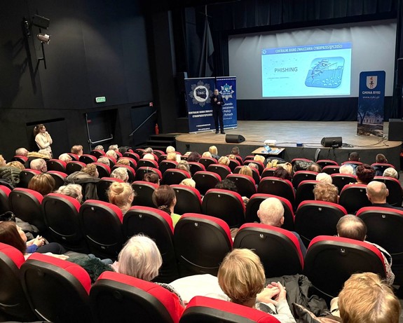 Spotkanie profilaktyczne w sali kina policjanta z seniorami. Funkcjonariusz stoi na scenie seniorzy siedzą w fotelach