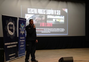 Spotkanie profilaktyczne w sali kina policjanta z seniorami. Funkcjonariusz stoi na scenie seniorzy siedzą w fotelach