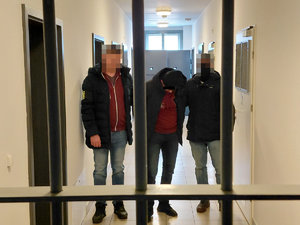 Dwóch policjantów pionu kryminalnego prowadzi zatrzymanego mężczyznę na korytarzu. Na pierwszym planie widoczna jest krata.