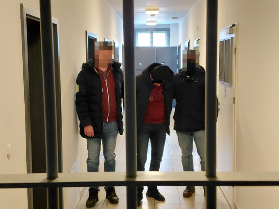 Dwóch policjantów pionu kryminalnego prowadzi zatrzymanego mężczyznę na korytarzu. Na pierwszym planie widoczna jest krata.